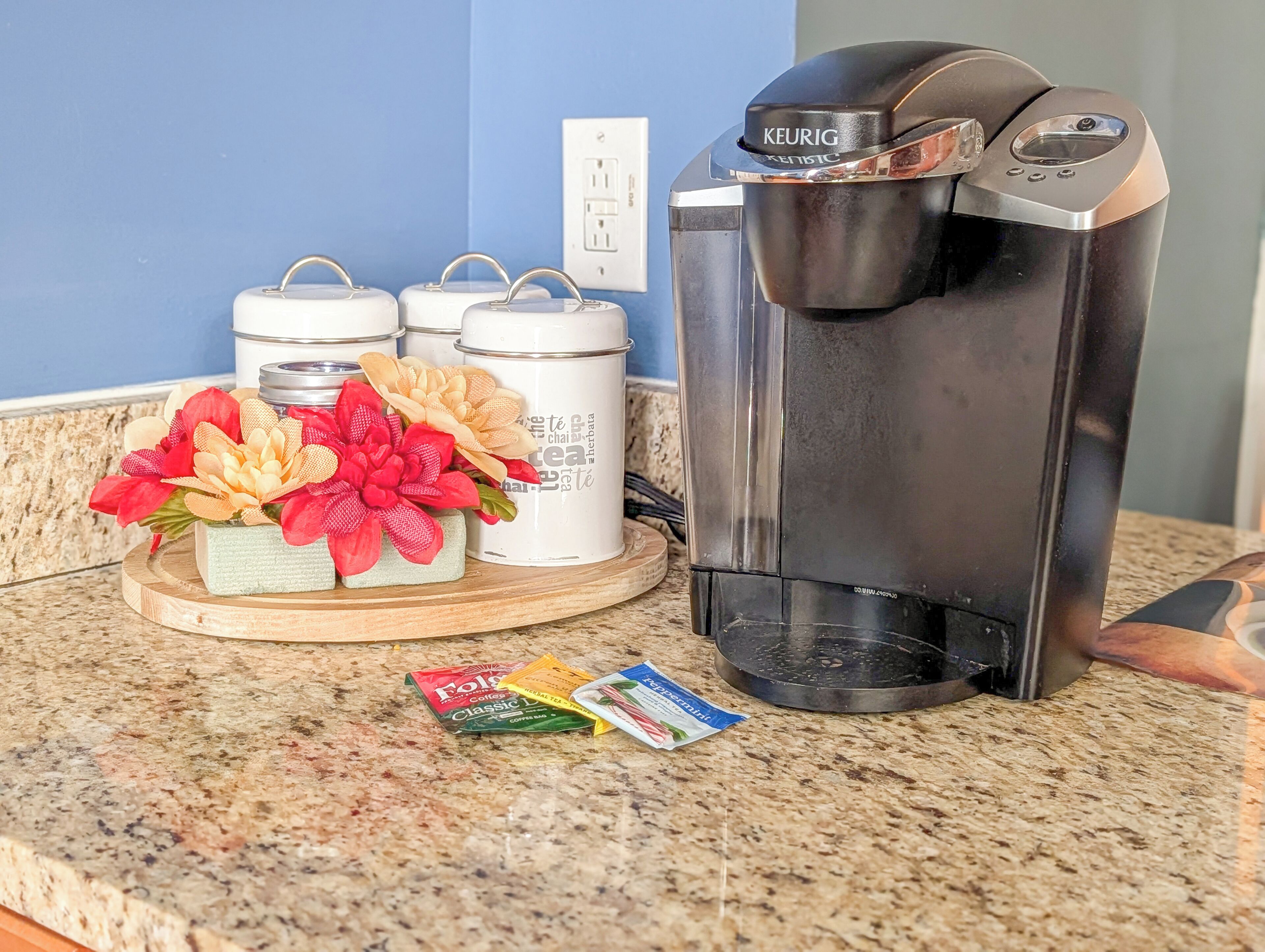 A coffee maker, three white containers, and a tray with artificial flowers on a kitchen counter.