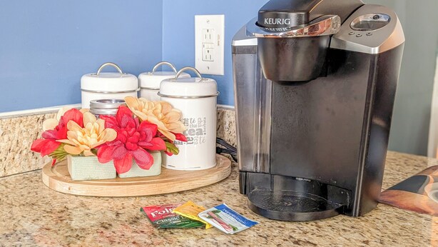Uma cafeteira, três recipientes brancos e uma bandeja com flores artificiais em uma bancada de cozinha.