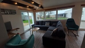 A modern living room with a dark-colored sofa, a teal armchair, and a small round table. There are large windows providing a view of the outdoors. - Comfortable house (10 persons), 1,5 km from the sea (Finistère Sud - Bretagne) (Plozévet)