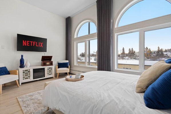 A modern bedroom with a large bed, a TV mounted on the wall, and a window with a view of a snowy landscape. - Upscale Historic Loft | TOP FLOOR | Downtown | Free Parking (Calgary)