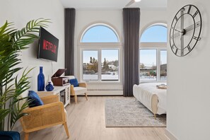 Ett modernt hotellrum med ett stort fönster, en väggklocka, en platt-TV som visar ”NETFLIX”, en säng, ett skrivbord med en bärbar dator, en stol och en krukväxt.
