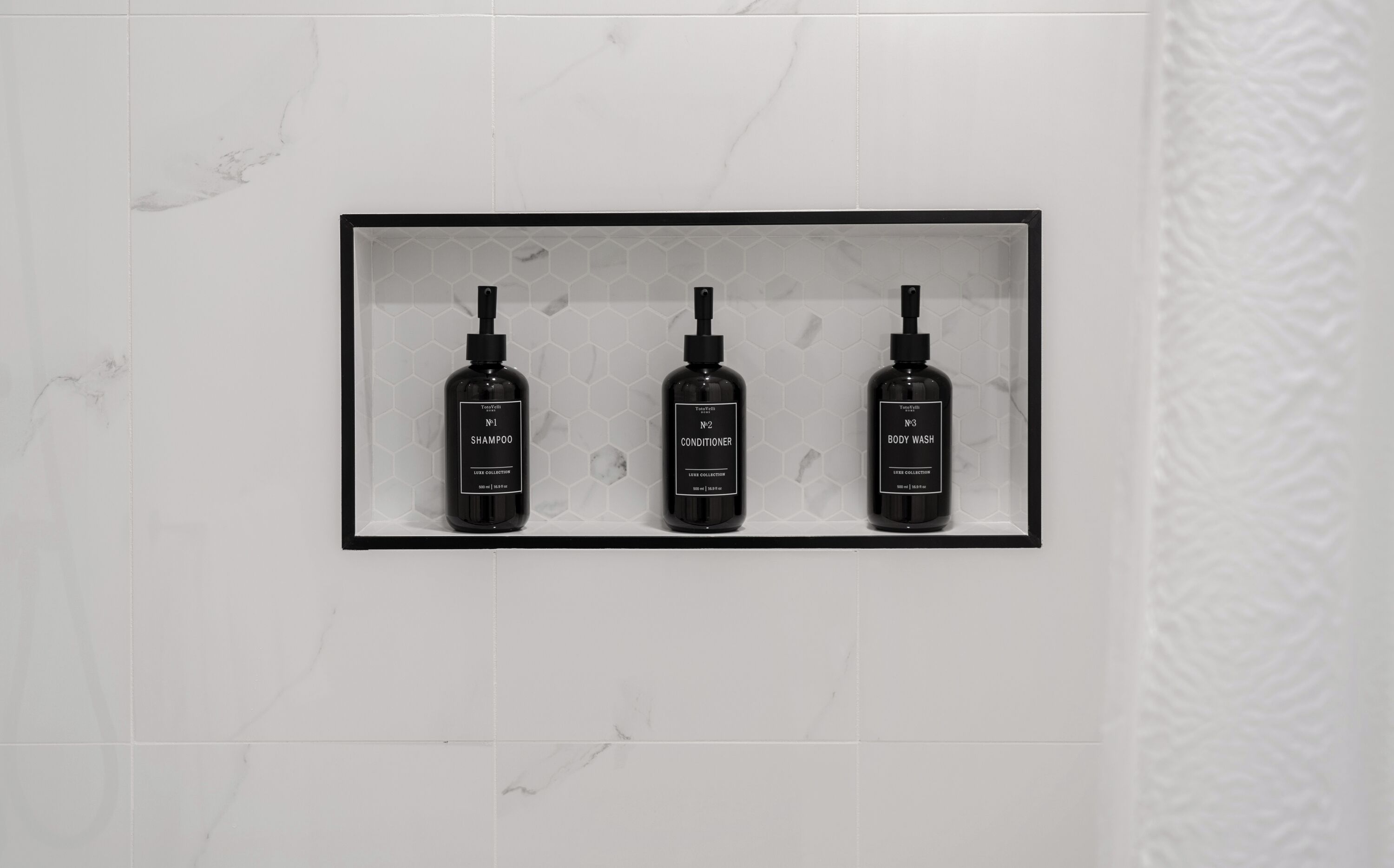 Ein Badezimmer mit Marmorwand und einem Regal, auf dem drei schwarze Flaschen mit den Aufschriften 'Shampoo', 'Conditioner' und 'Body Wash' stehen.