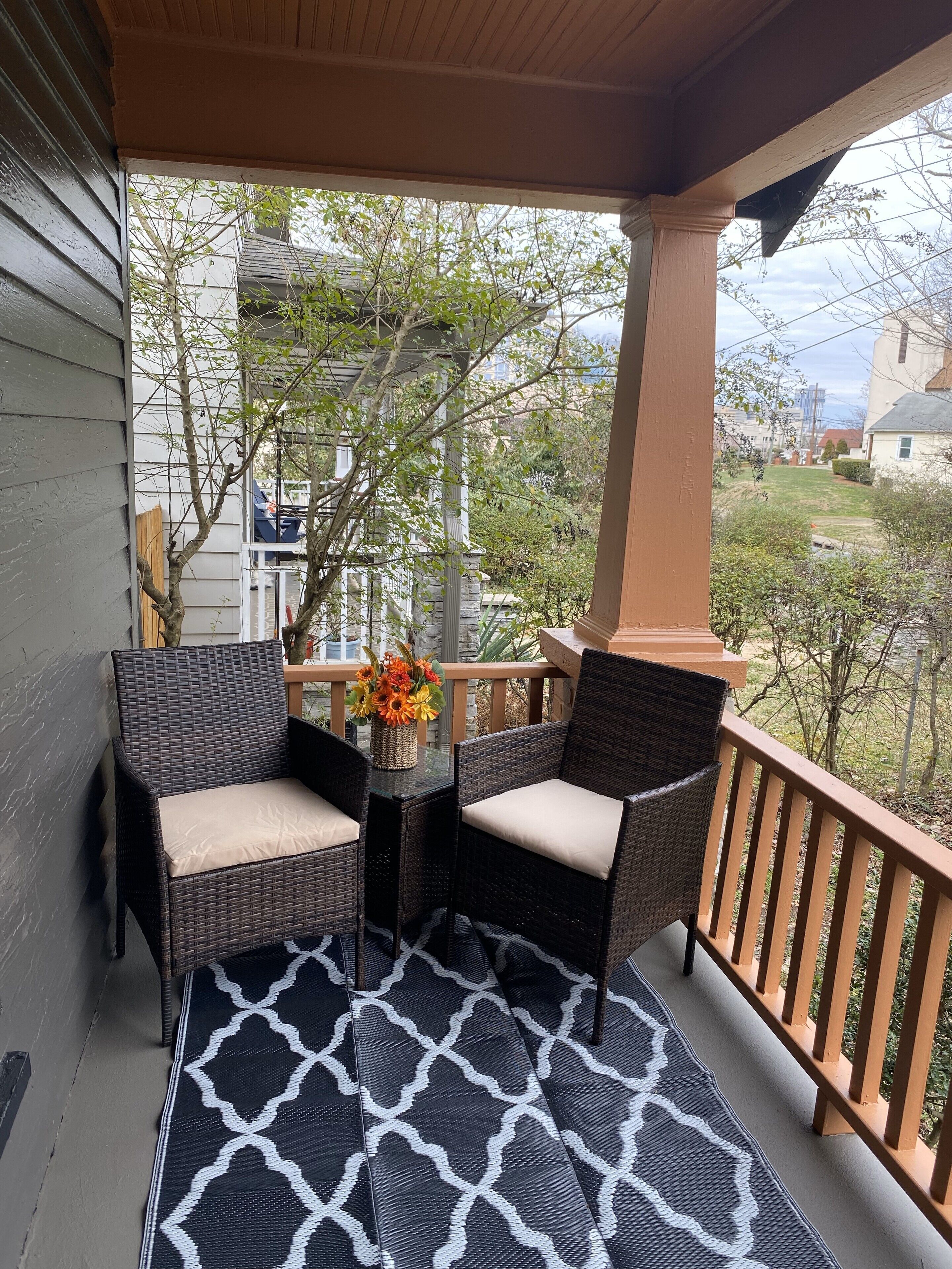 A covered porch with wicker chairs and a table, a patterned rug, and a view of a residential area.