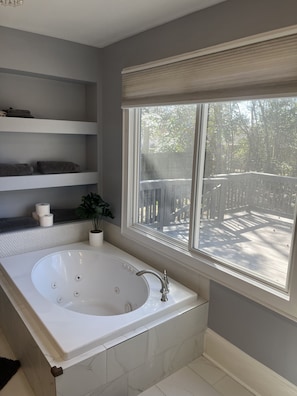Un baño con una bañera grande, estantes con toallas, una planta en maceta y una ventana con vista a una terraza.