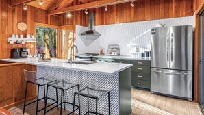 Cocina moderna con paredes de madera, una isla grande con taburetes de bar, un refrigerador de acero inoxidable y un ventanal con vista a árboles.