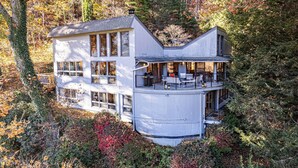 Una casa moderna de dos pisos con amplios ventanales y un balcón, rodeada de un bosque denso.