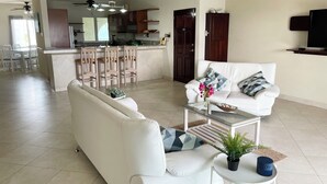 Una sala de estar moderna con un sofá blanco, una mesa de centro de vidrio y un televisor sobre un mueble. Hay una zona de cocina con armarios de madera y un comedor con sillas.
