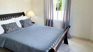 Una cama bien tendida con una colcha azul y almohadas estampadas, una lámpara de noche y una ventana con cortinas.