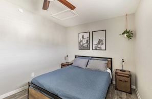 Un dormitorio con una cama, mesitas de noche, un ventilador de techo y dos cuadros en la pared.