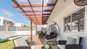 Une terrasse extérieure couverte, aménagée avec des meubles en osier, un fauteuil suspendu et une piscine.