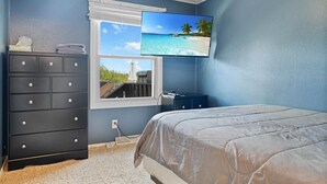 A bedroom with a bed, a dresser, a window with a view of a beach, and a television mounted on the wall.