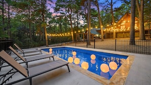 Una piscina con sillones de descanso e iluminacion de guirnaldas, rodeada de árboles y una cabaña de madera.