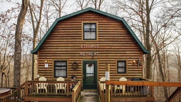 A log cabin with a green door, labeled "ETHAN'S RETREAT," featuring a porch with chairs and a small table.