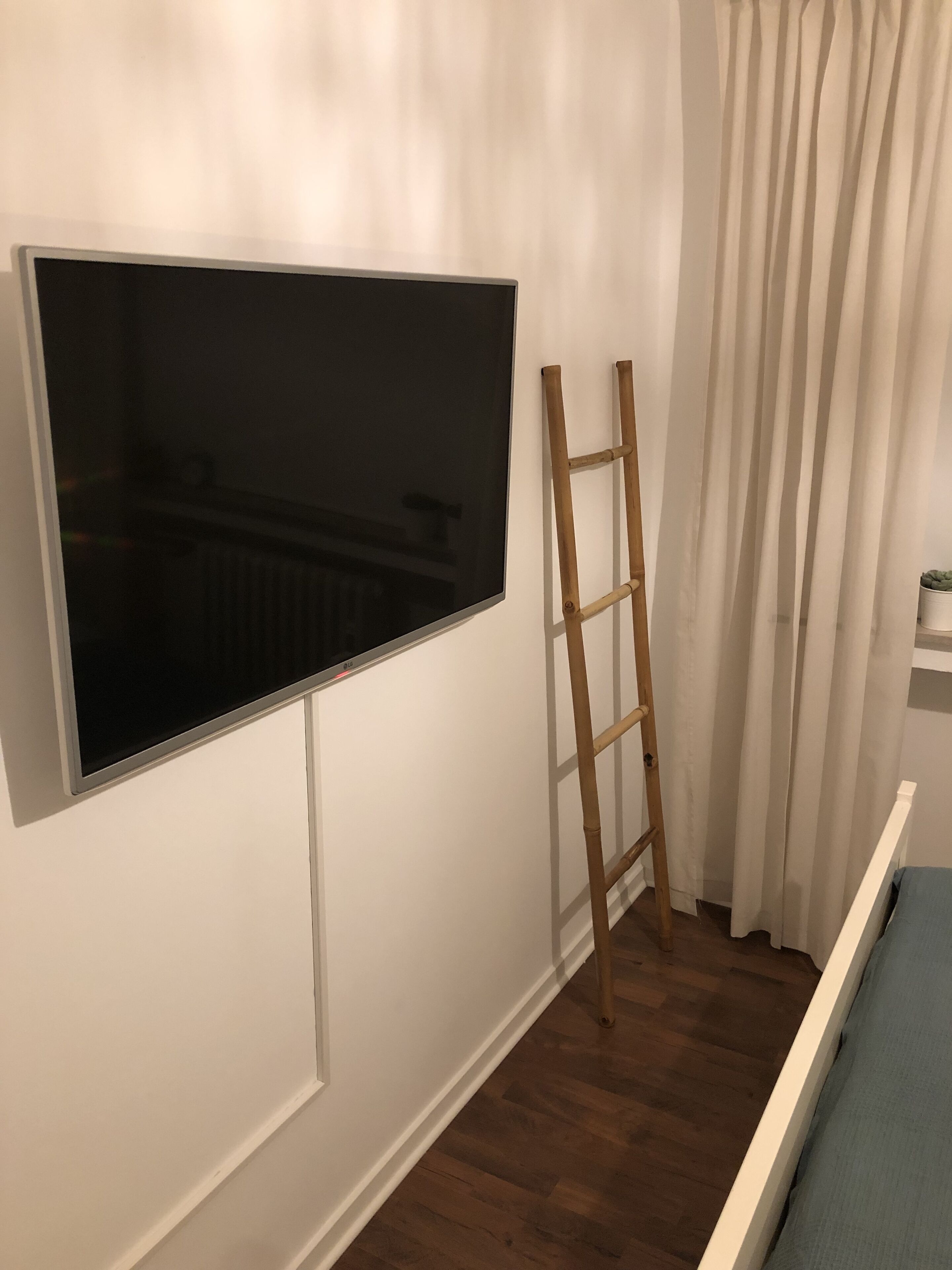 A room with a large flat-screen TV mounted on the wall, a wooden ladder leaning against the wall, and a bed with a blue mattress.