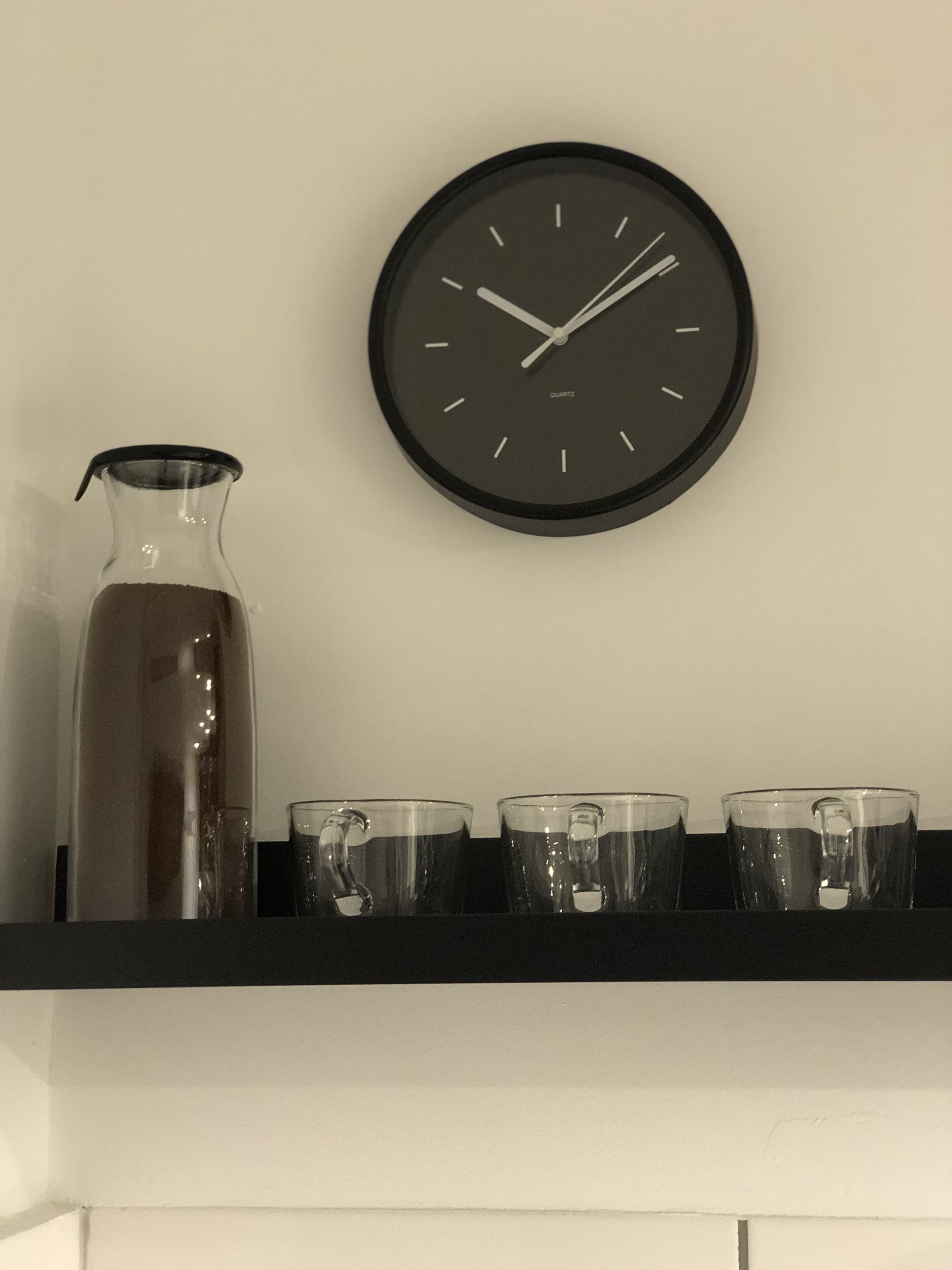A kitchen with a black clock, a glass pitcher, and three clear cups on a black shelf.
