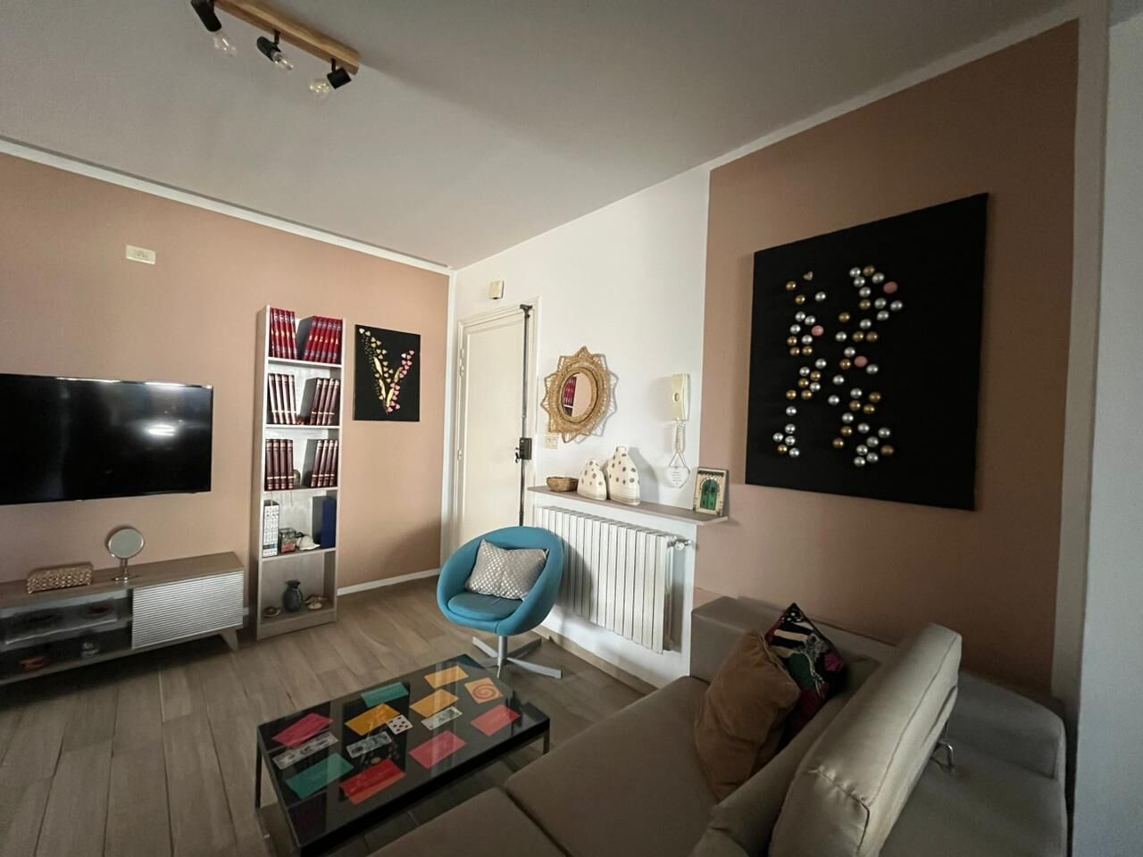 A living room with a flat-screen TV, a coffee table, a blue armchair, a sofa, and a bookshelf.