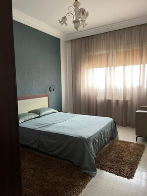 A bedroom with a bed, a chandelier, a window with curtains, and a rug. - Apartment tourist area corniche Sousse (Sousse)