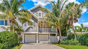 A two-story house with a garage, surrounded by lush greenery and palm trees. - Prime Holmes Beach Location | Heated Pool | Walk to the Beach (Holmes Beach)