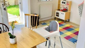A bright, open-plan living space with a wooden dining table, chairs, a colorful rug, a kitchen area with a toy kitchen set, and a view of a garden through an open door. - Historic family house near Longleat (Warminster)
