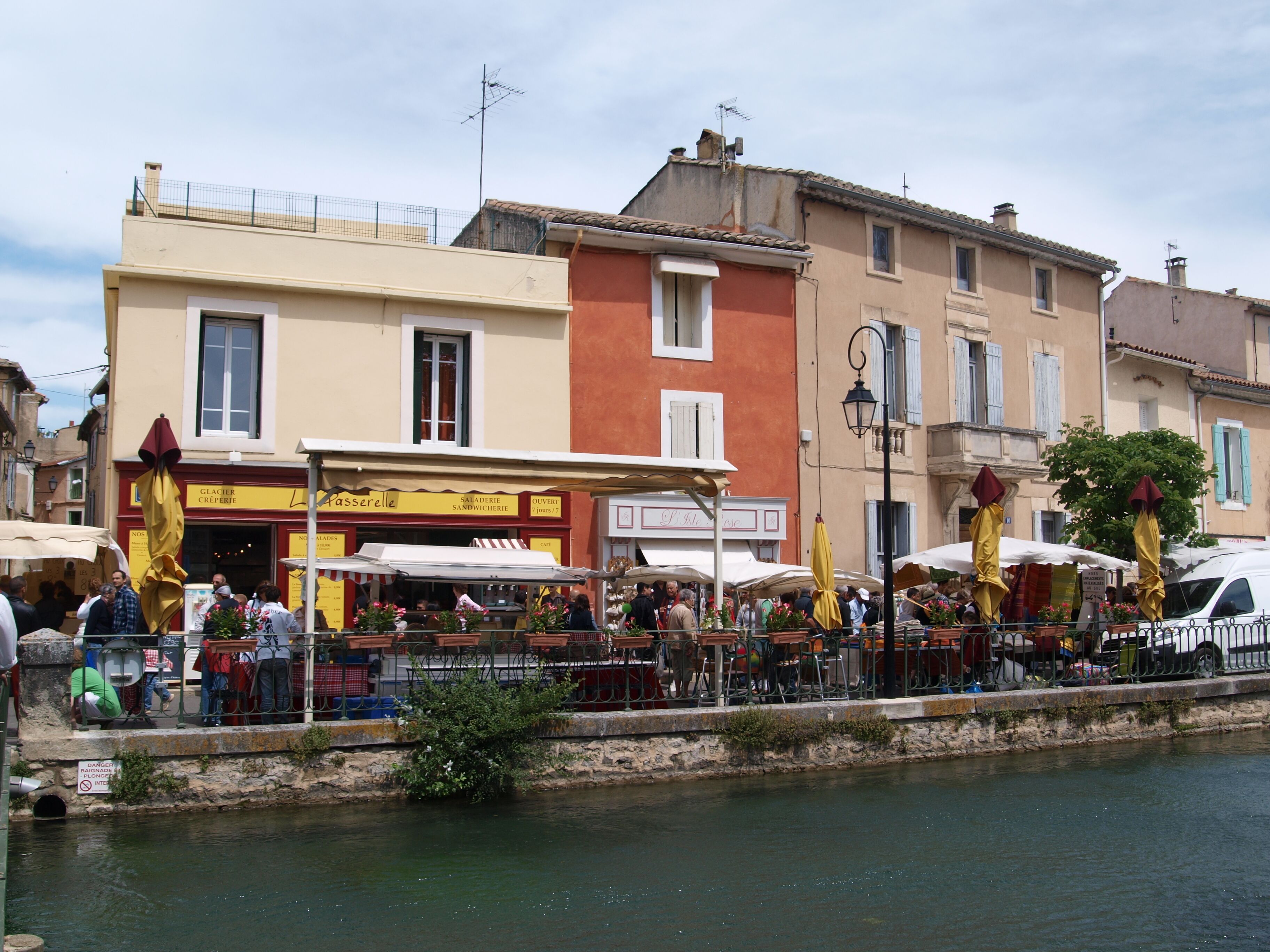 A riverside café with outdoor seating, a stone wall, and a building with a sign for a creperie.