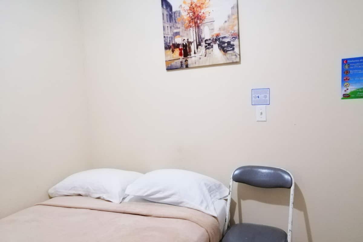 A bed with a beige blanket, a white pillow, and a grey chair against a wall with a painting and a notice.