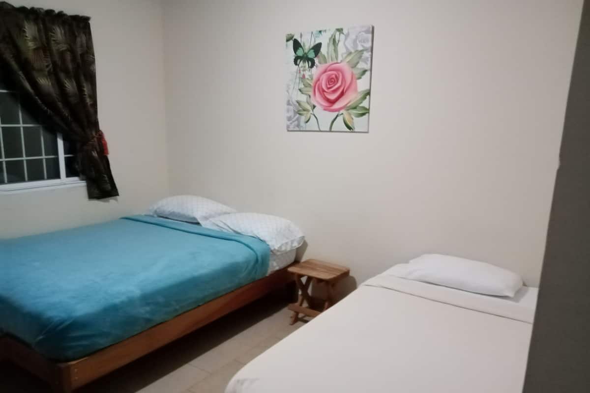 A bedroom with two beds, one with a blue bedspread and the other with a white bedspread. There is a small wooden nightstand between the beds. A window with curtains is visible on the left side of the room.
