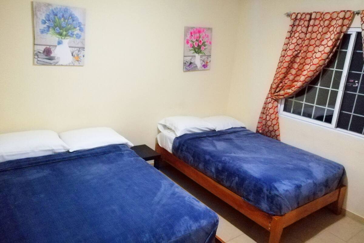 A room with two beds, each with a blue bedspread and white pillows. There are two paintings on the wall, one with blue flowers and another with pink flowers. A window with a patterned curtain is visible.