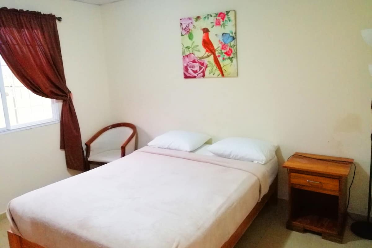 A bedroom with a bed, a chair, a nightstand, a window with curtains, and a floral painting on the wall.