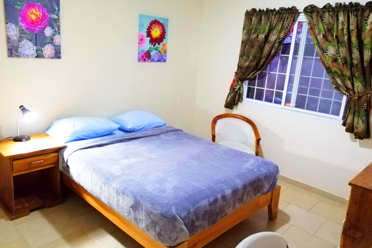 A bedroom with a bed, a nightstand, a chair, a window with curtains, and two wall-mounted flower paintings.