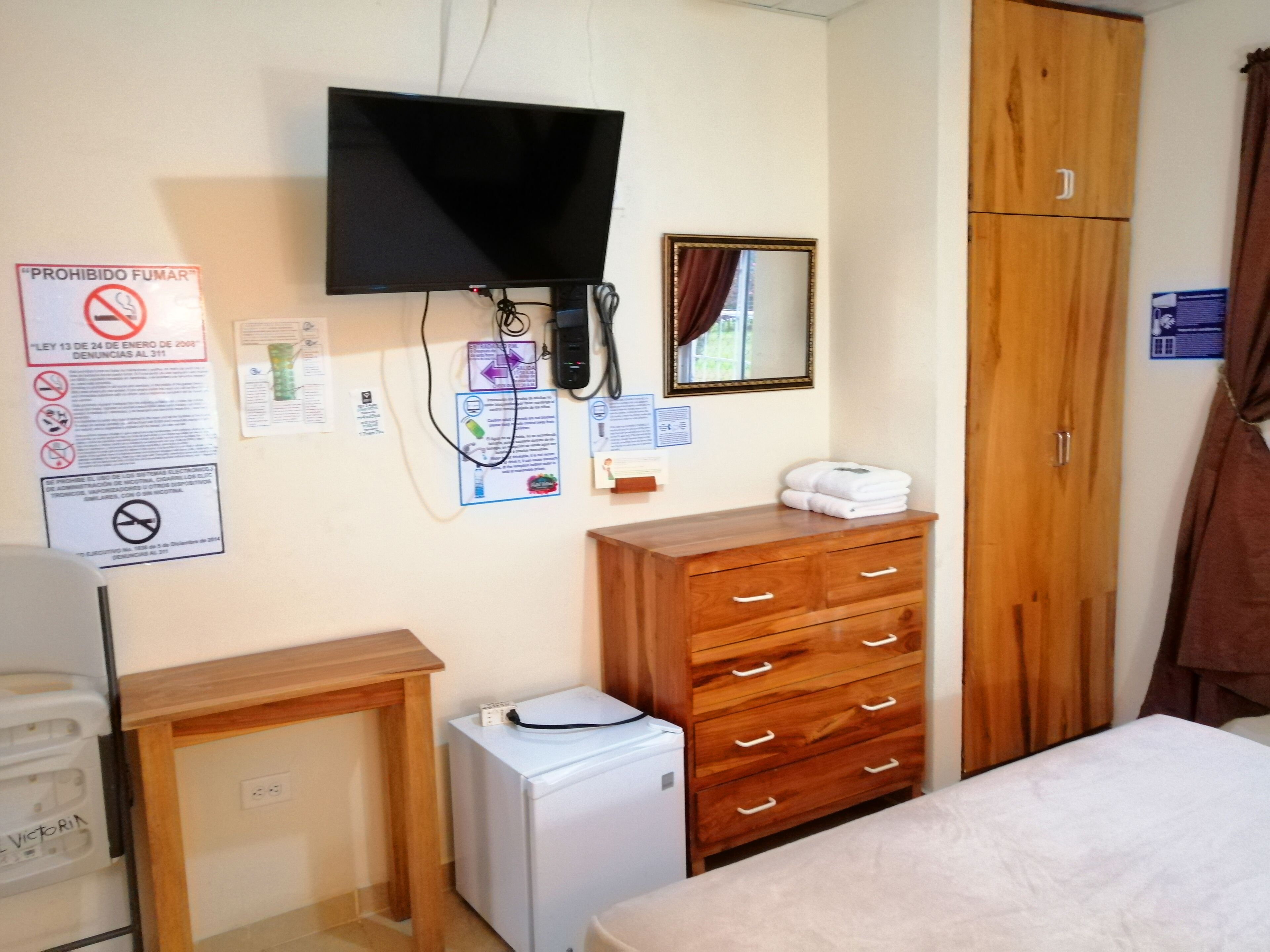 Un dormitorio con un aparador de madera, un refrigerador blanco pequeño, un televisor montado en la pared y una cama.
