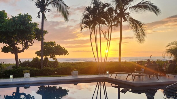 Ein Poolbereich mit Sonnenuntergangsblick, Palmen und einer Gruppe von Personen, die auf Liegestühlen sitzen.