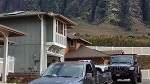 Un quartier résidentiel avec une maison et des véhicules garés, entouré de montagnes verdoyantes et de nuages.