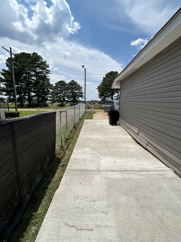 A concrete pathway leading to a building with a metal fence on one side and a grassy area with trees on the other.