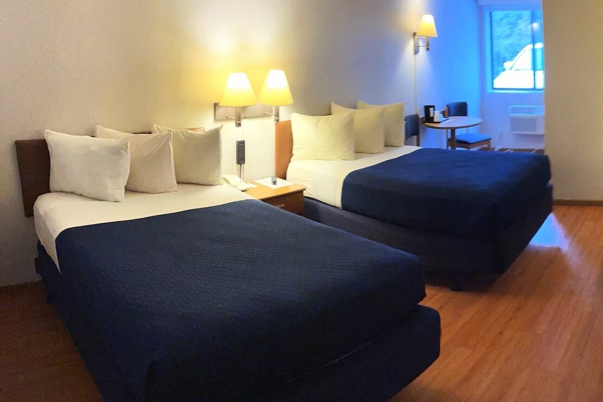 Ein Hotelzimmer mit zwei Betten, einem Schreibtisch und einem Sessel. Wandlampen und ein Fenster mit Blick auf ein Auto.
