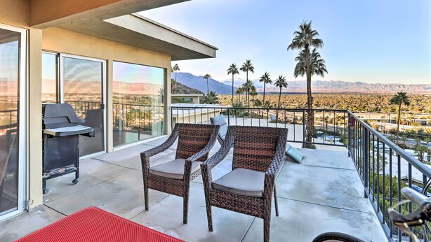 A balcony with wicker chairs, a grill, and a view of palm trees and mountains.