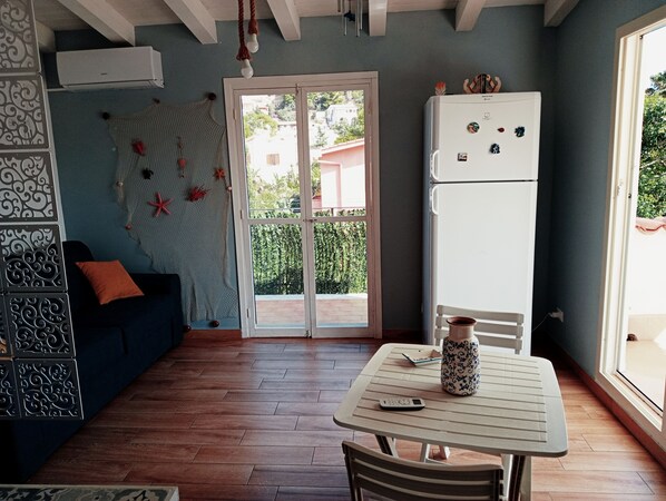 A compact kitchen with a white refrigerator, a dining table with a vase, and a blue sofa with orange cushions. - Apartment with sea view (Santa Flavia)
