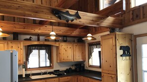 Une cuisine avec des meubles en bois, un poisson décoratif fixé au plafond, des luminaires suspendus et une fenêtre ornée d’un rideau à carreaux.