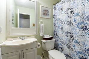 Un baño con lavamanos blanco, inodoro y cortina de ducha con motivos tropicales.