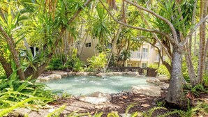 A natural pool surrounded by lush greenery and trees, with a building in the background.