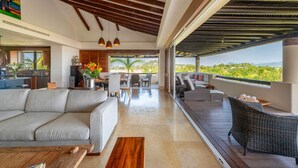 A modern living room with a sofa, dining table, and chairs, a kitchen area, and a balcony with a view. - PH Vista Encanto in El Encanto by Mita Residential. Golf & Ocean View, Pool, Golf Cart (Punta Mita)