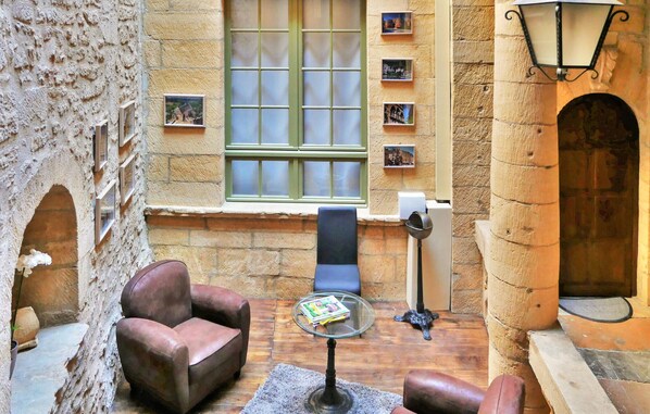 A cozy room with a stone wall, a window with green shutters, a chair, a small table with a magazine, and a door with a doormat.