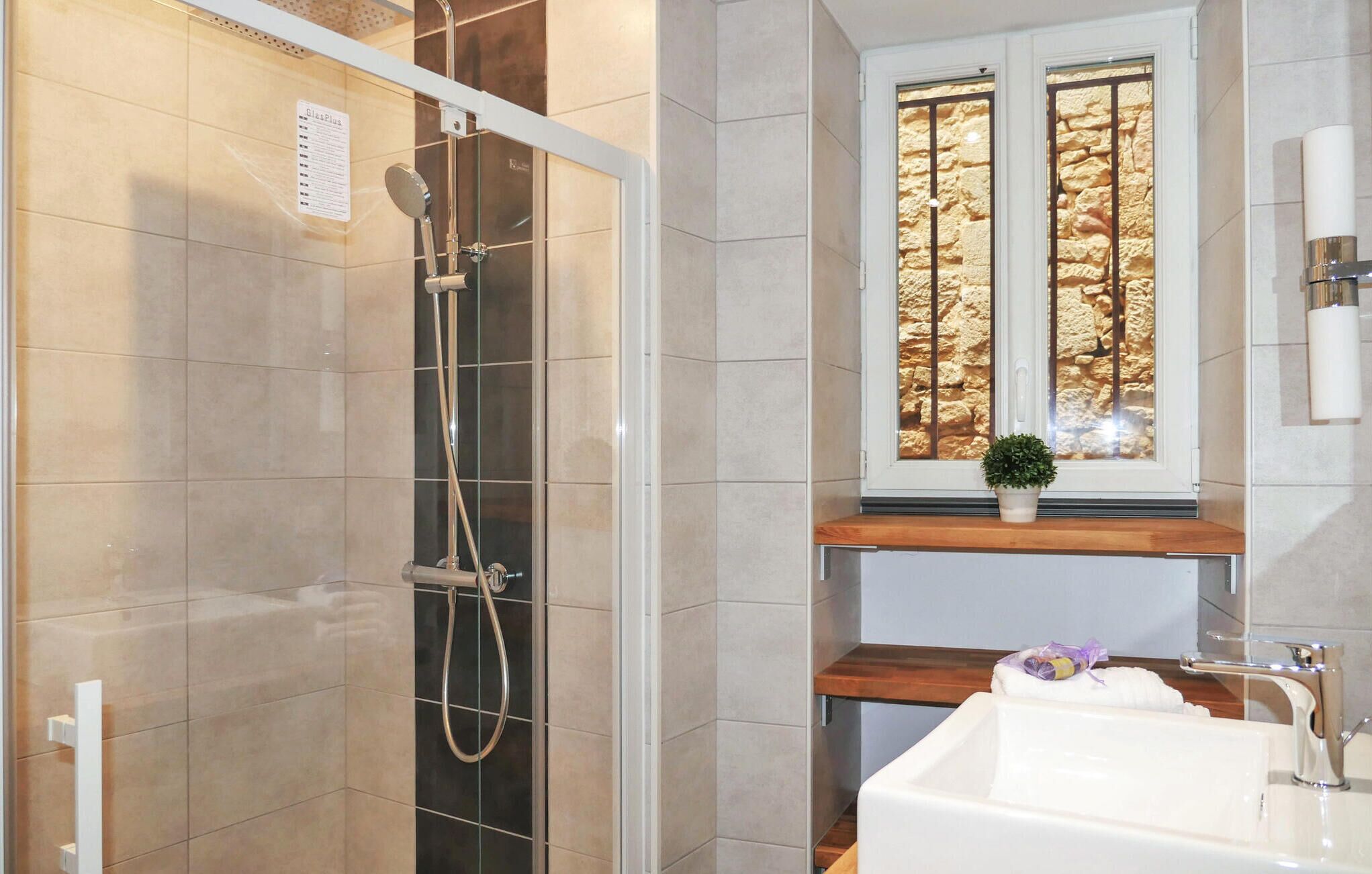 Une salle de bain moderne avec une douche en verre, une fenêtre dotée d’une façade en pierre et une étagère en bois sur laquelle est posée une plante en pot.