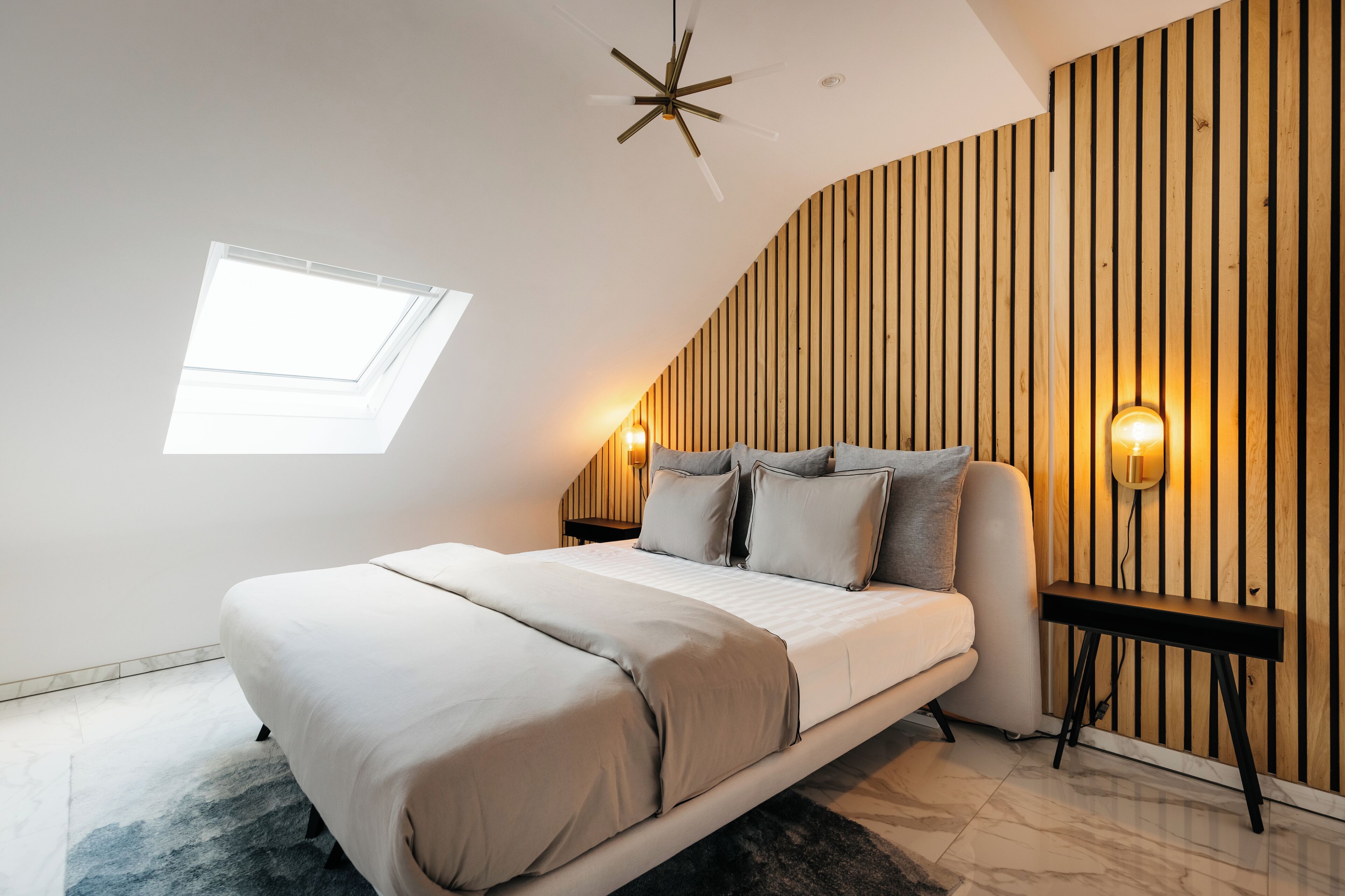 Un dormitorio moderno con techo inclinado, tragaluces, cama con colcha color beige, dos lámparas de noche y paneles de madera.