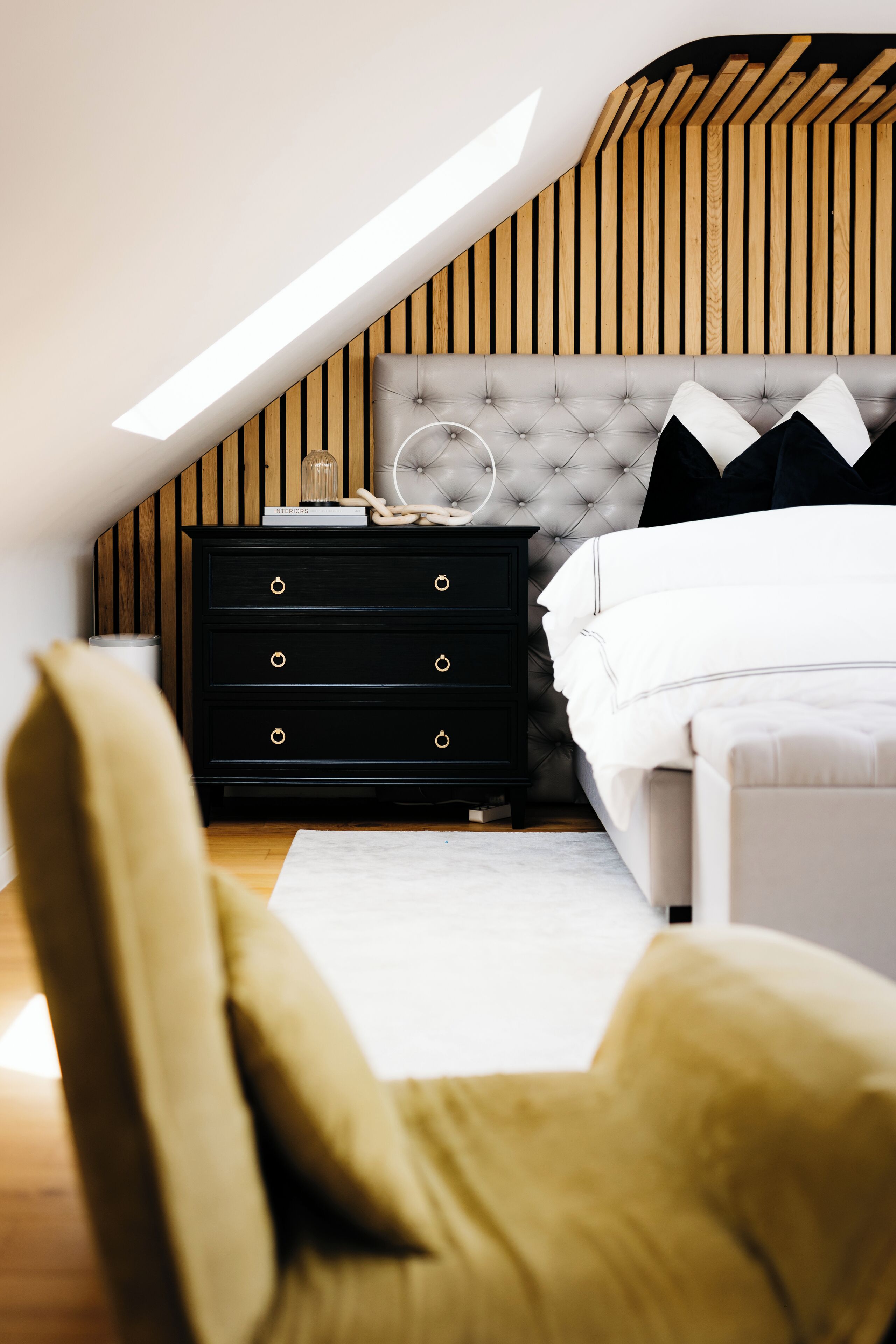 A bedroom with a sloped ceiling, a tufted headboard, a black dresser with gold handles, a white bed, and a yellow armchair.
