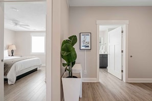 A hotel room with a bed, a nightstand, a lamp, a large potted plant, a framed picture on the wall, and a bathroom visible through an open door. - 🌆 539 Bay St Lakeview Condo | Walk to Downtown & Beach (Traverse City)