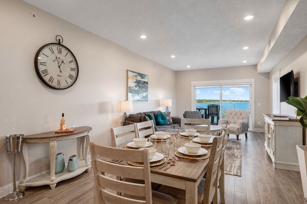 A dining area with a wooden table set for four, a large wall clock, and a view of the outdoors. - 🌆 539 Bay St Lakeview Condo | Walk to Downtown & Beach (Traverse City)