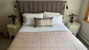 A neatly made bed with a quilted beige cover, two white pillows, and a patterned throw pillow. There are two bedside tables with lamps, a waste basket, and a window with curtains. - Beautiful Modern Condo Downtown/Convention Center (Seattle)