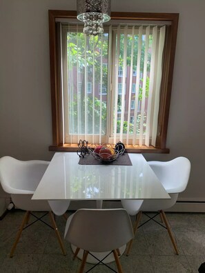 Enjoy meals in this dining area with a table, chairs, and a chandelier, offering a view of the surrounding building and greenery.