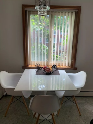 Enjoy meals in this dining area with a table, chairs, and a chandelier, offering a view of the surrounding building and greenery.