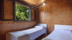 A wooden cabin room with two beds, a window with a view of trees, and a wall-mounted light fixture.
